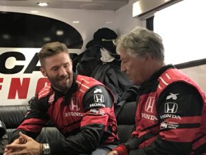 Two men in red and black jackets sitting on a car.